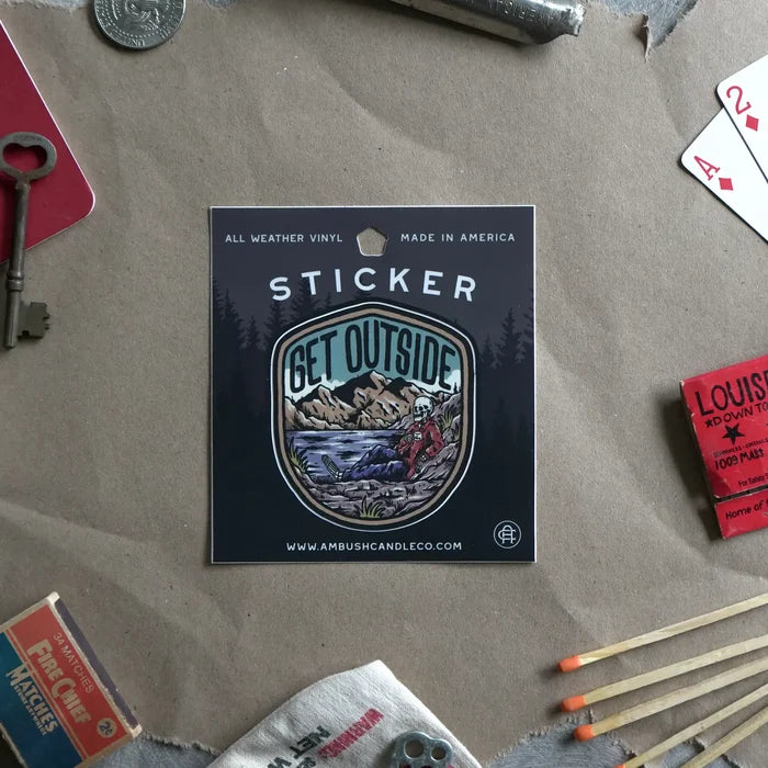 The Ambush Candle Co Get Outside Sticker, featuring the "GET OUTSIDE" text and a mountain scene, is placed among fallen autumn leaves. This weatherproof sticker, made in the USA, can be removed without damage.