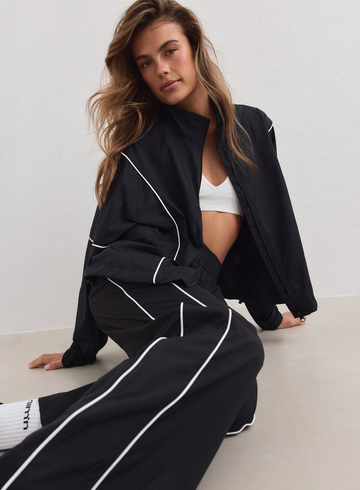 Musta aim’n Balance Windstopper Tracksuit Jacket valkoisilla tereillä istuvan mallin yllä.