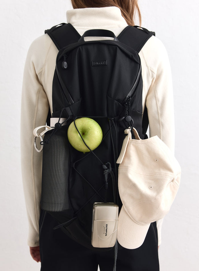 Musta aim'n Outdoor Daypack -reppu, johon on kiinnitetty vihreä omena, juomapullo, lippis ja kamera.
