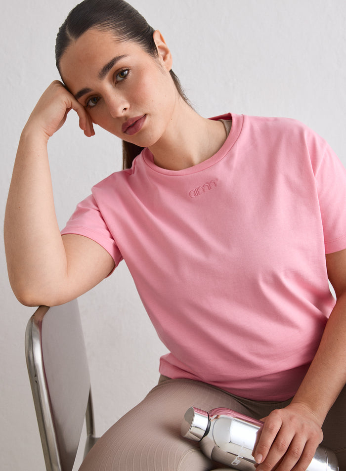 Nainen yllään Blush Boxy T-shirt, jossa on brodeerattu aim'n-logo, istumassa tuolilla ja pitelemässä hopeista vesipulloa [L-174cm]