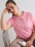 Nainen yllään Blush Boxy T-shirt, jossa on brodeerattu aim'n-logo, istumassa tuolilla ja pitelemässä hopeista vesipulloa [L-174cm]