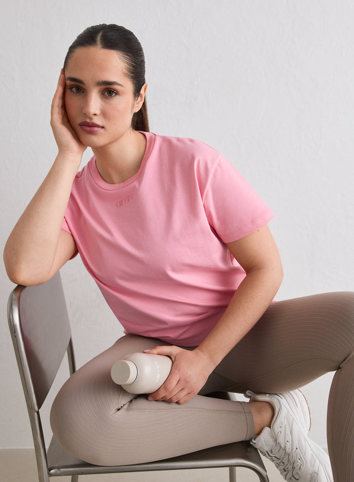 Nainen istuu tuolilla yllään Blush Boxy T-shirt, jossa on brodeerattu aim'n-logo [L-174cm]