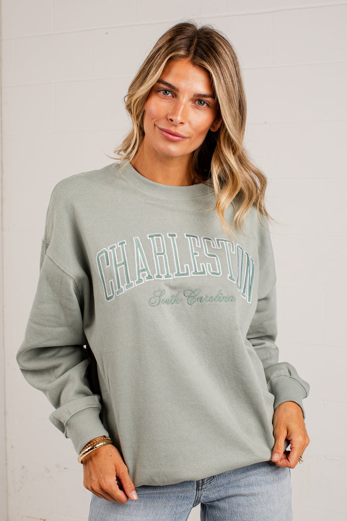 A woman wearing a sage green sweatshirt with 'CHARLESTON South Carolina' printed in arched teal and white lettering across the chest, paired with light blue jeans, set against a white paneled wall.