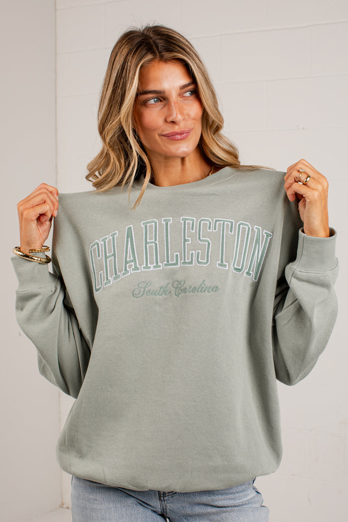 A woman wearing a sage green sweatshirt with 'CHARLESTON South Carolina' printed in teal on the front, holding the sleeves out to show the fit against a white brick wall.