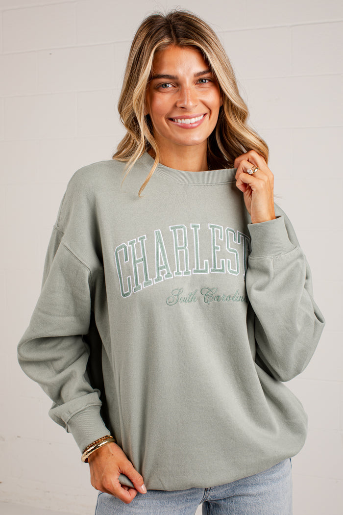 A woman wearing a sage green sweatshirt with 'CHARLESTON South Carolina' printed in light blue and green on the front, paired with light blue jeans, standing against a white wood-paneled wall.