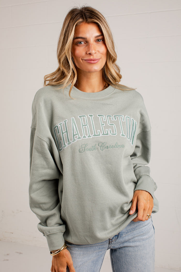 A woman wearing a sage green crewneck sweatshirt with 'CHARLESTON South Carolina' printed in teal across the chest, paired with light blue jeans, set against a white brick wall background.