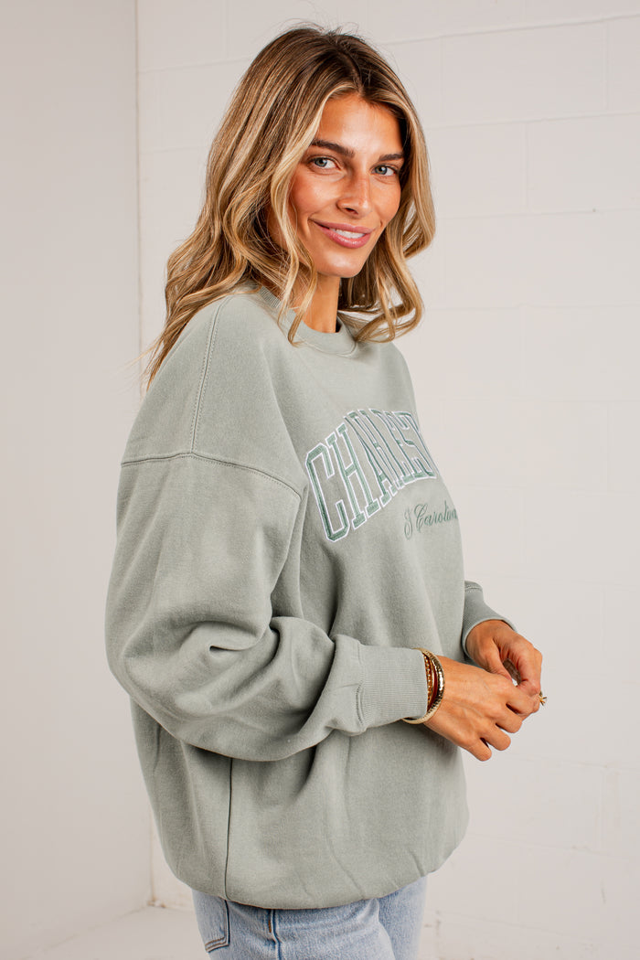 A woman wearing a sage green crewneck sweatshirt with 'CHICAGO' printed in light blue arched lettering across the chest, paired with light blue jeans, standing against a white brick wall.