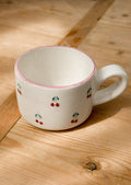 A cream ceramic mug with hand painted cherry motif and pink top rim.