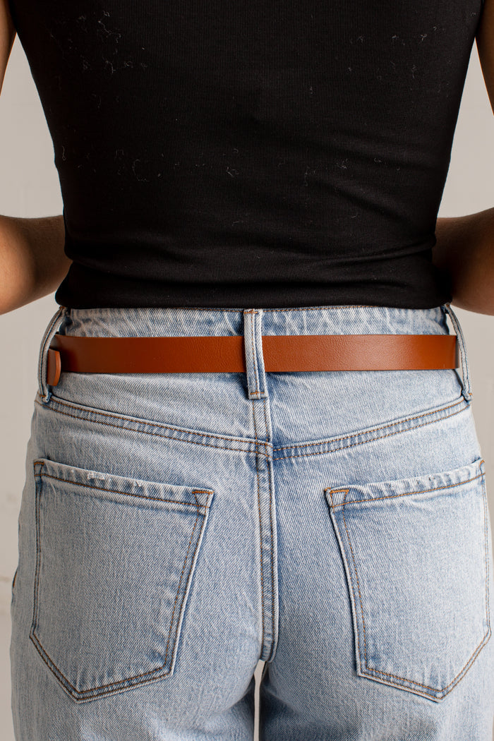 A close-up back view of a person wearing a slim brown leather belt with a small gold-tone accent on the left side, paired with light-wash blue jeans and a black ribbed top.