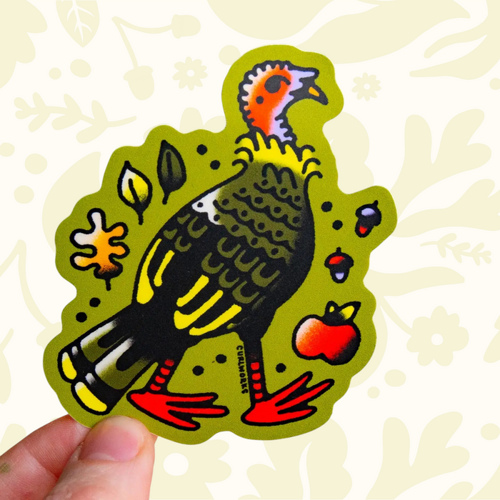 Hand holding Curlworks Turkey Vinyl Sticker with red feet, fall leaves, berries, and an apple on green background. Perfect decor.