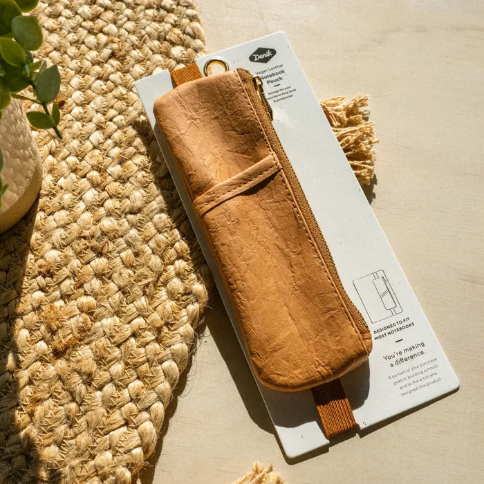 Denik Caramel Crinkle Vegan Leather Notebook Pouch displayed on its box by a woven mat and potted plant, stylish desk setup.