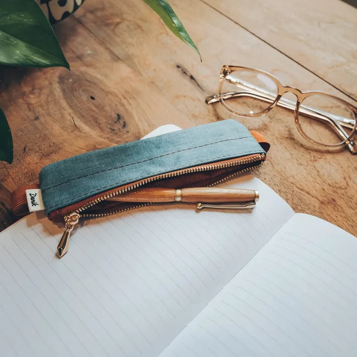 Denik Denim Distressed Blue Notebook Pouch, open notebook, pen, glasses, and green leaf rest on a wooden table.