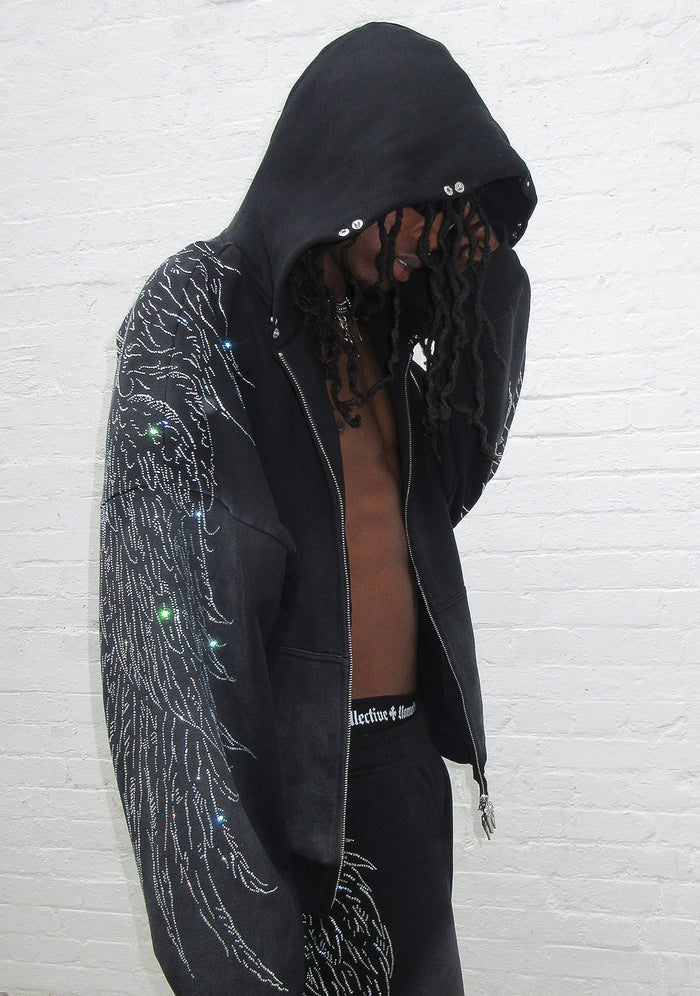 Model wearing a hoodie in washed black with rhinestone wing design and hood up, showcasing front details.