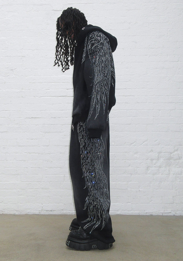 Model wearing fallen rhinestone oversized sweatpants washed black with rhinestone wing design, side view.