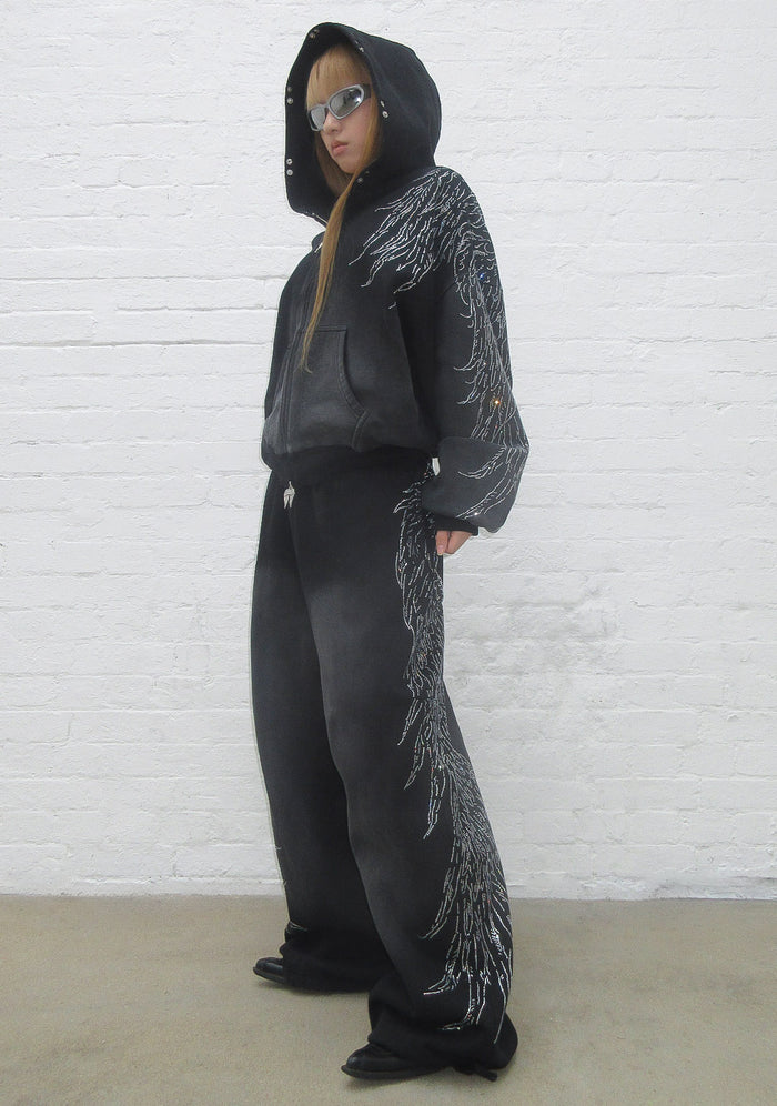 Model wearing black fallen rhinestone oversized sweatpants with large rhinestone wing design and pockets.