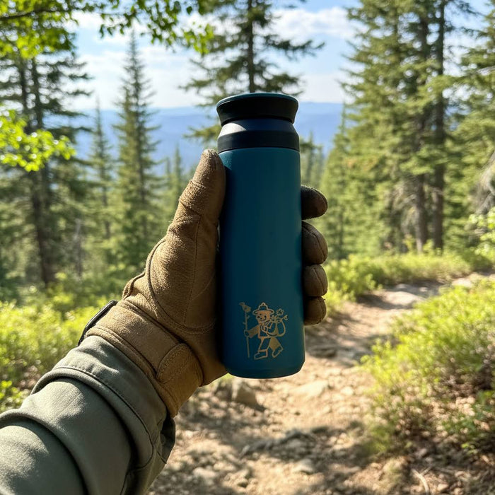 Hand in glove holds Forager Provisions Betta Fish Blue 500ml flask on forest trail with scenic trees and mountains backdrop.