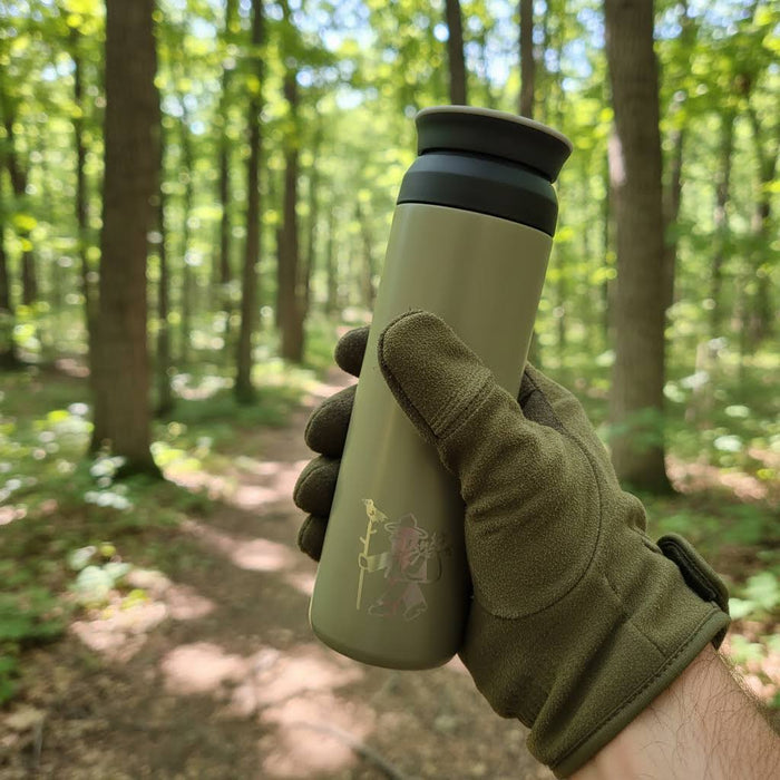 Gloved hand holding Forager Provisions Mossy Stone 500ml stainless steel flask outdoors on sunlit forest hiking trail.