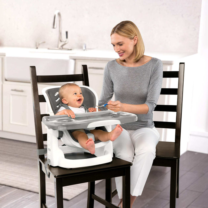 Ingenuity SmartClean ChairMate portable high chair and toddler booster seat in slate attached to dining chair with removable wipeable tray and safety straps