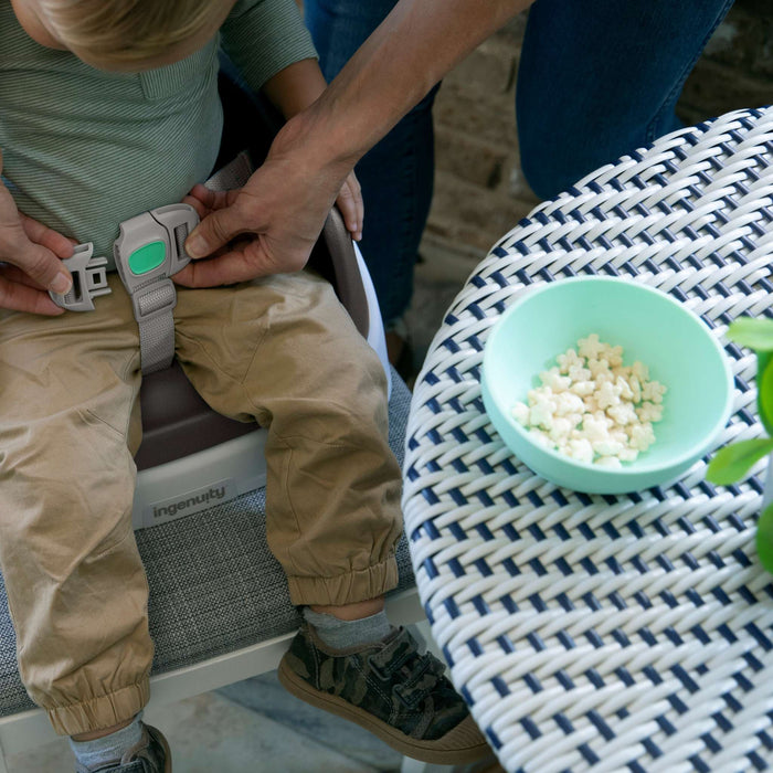 Ingenuity SmartClean Toddler Booster Seat in slate with 3-point harness and washable foam insert attached to dining chair