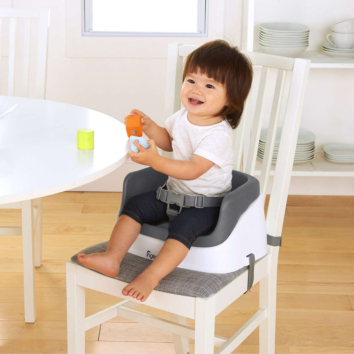 Ingenuity SmartClean Toddler Booster Seat in slate color with removable foam insert and 3-point harness attached to dining chair