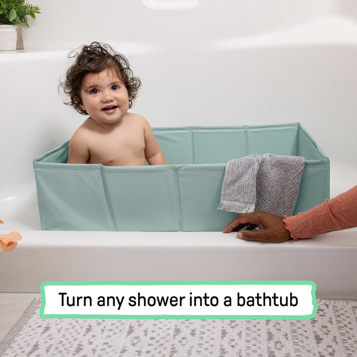 Baby sitting in an unfolded Ingenuity Tuckaway Travel Tub set inside a bathtub with a towel hanging on the side and an adult's hand nearby