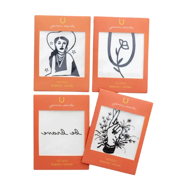 Four non-toxic temporary tattoos packaged in an orange envelope include designs of a woman with a hat, a rose, the words "go brave", and an intricate floral hand with leaves. Perfect for adding western vibes to any look, these Jenni Earle Cowgirl Dreamin' Temporary Tattoos bring a touch of adventure wherever you go.