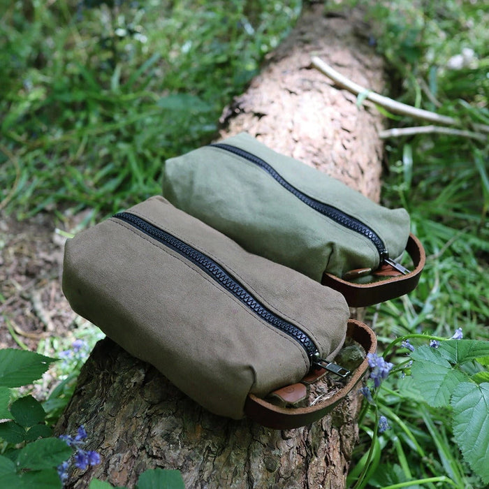 Forager Provisions LTD green and brown canvas toiletry bags with YKK zippers resting on a log in a forest setting.