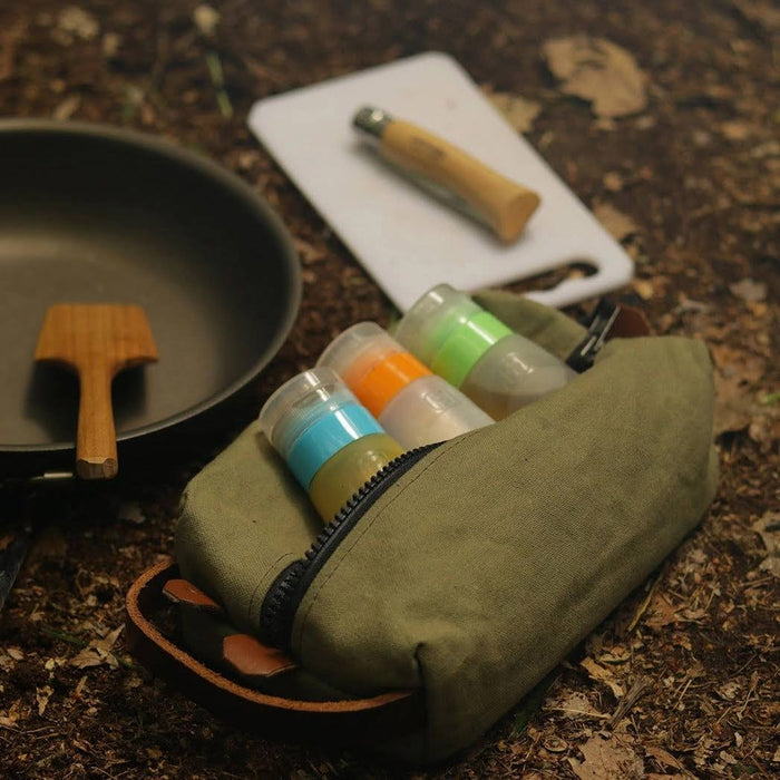Forager Provisions LTD green canvas toiletry bag filled with travel bottles, sitting on forest ground near camping gear.
