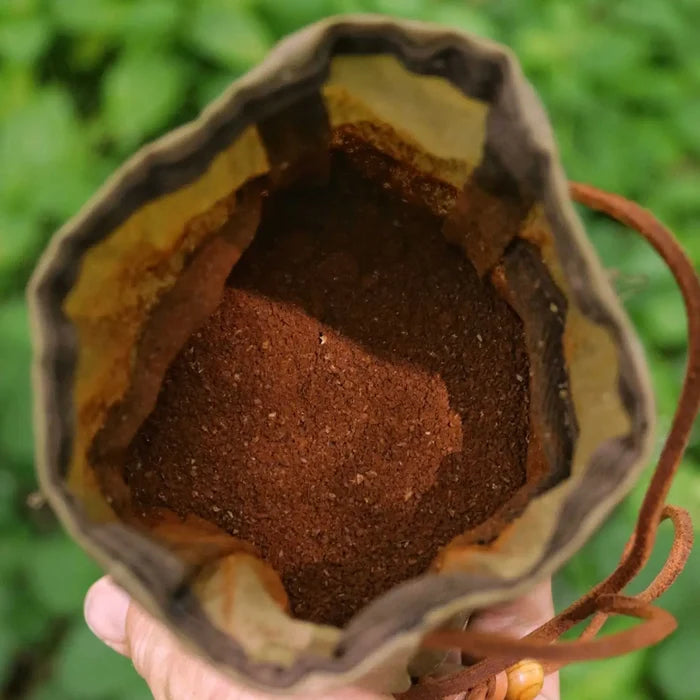Hand holding Journeyman Craft khaki green waxed canvas coffee pouch outdoors, blurred foliage in background. Eco travel gear.
