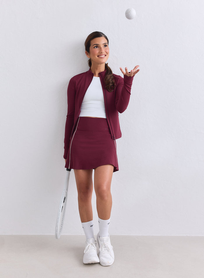Nainen yllään merlot-värinen aim'n Sense Zip Jacket ja yhteensopiva hame heittämässä tennispalloa. [XS-165cm]