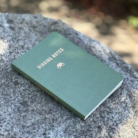 Birding Notes notebook by The Mountaineers Books with binoculars on gray rock outdoors—birdwatching journal in nature.