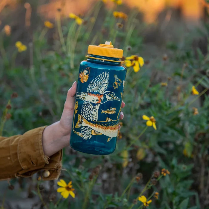 Hand holding Mustard Beetle 32oz Nalgene Malibu Creek water bottle for Cal Trout, with wildflowers outdoors.
