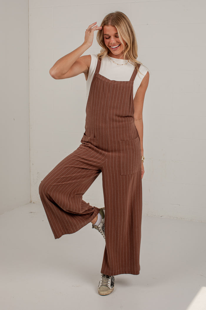 A woman modeling a brown pinstriped wide-leg jumpsuit over a white sleeveless top, standing in a bright studio setting.