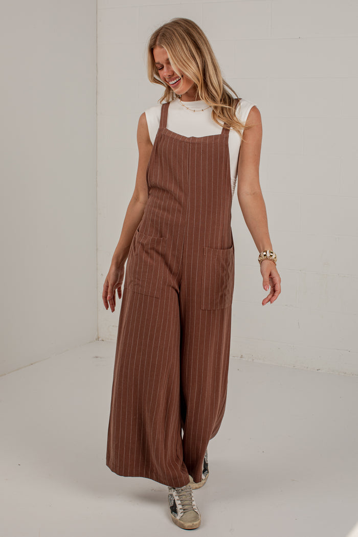 A woman modeling brown corduroy pinstriped overall jumpsuit with wide legs, worn over a white sleeveless top, in a white studio setting.
