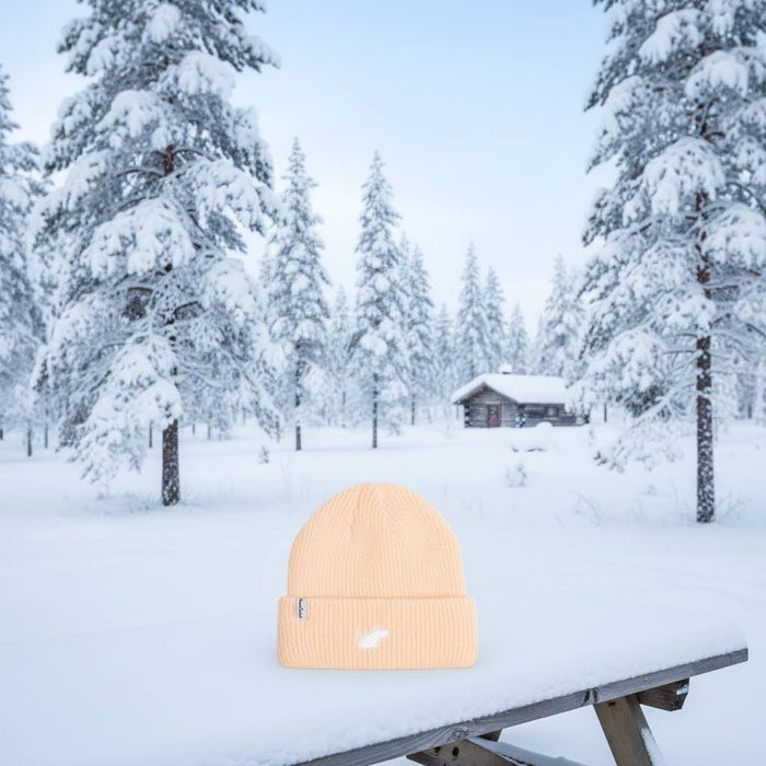 Peach Moomin Classics Winter Beanie by Nordicbuddies on snowy table, trees and cabin in wintery background, cozy scene.