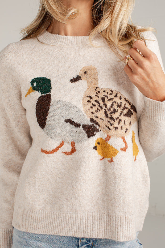 A woman wearing a cream-colored knit sweater with a detailed knitted duck family motif on the front, featuring a mallard, a speckled duck, and two yellow chicks.