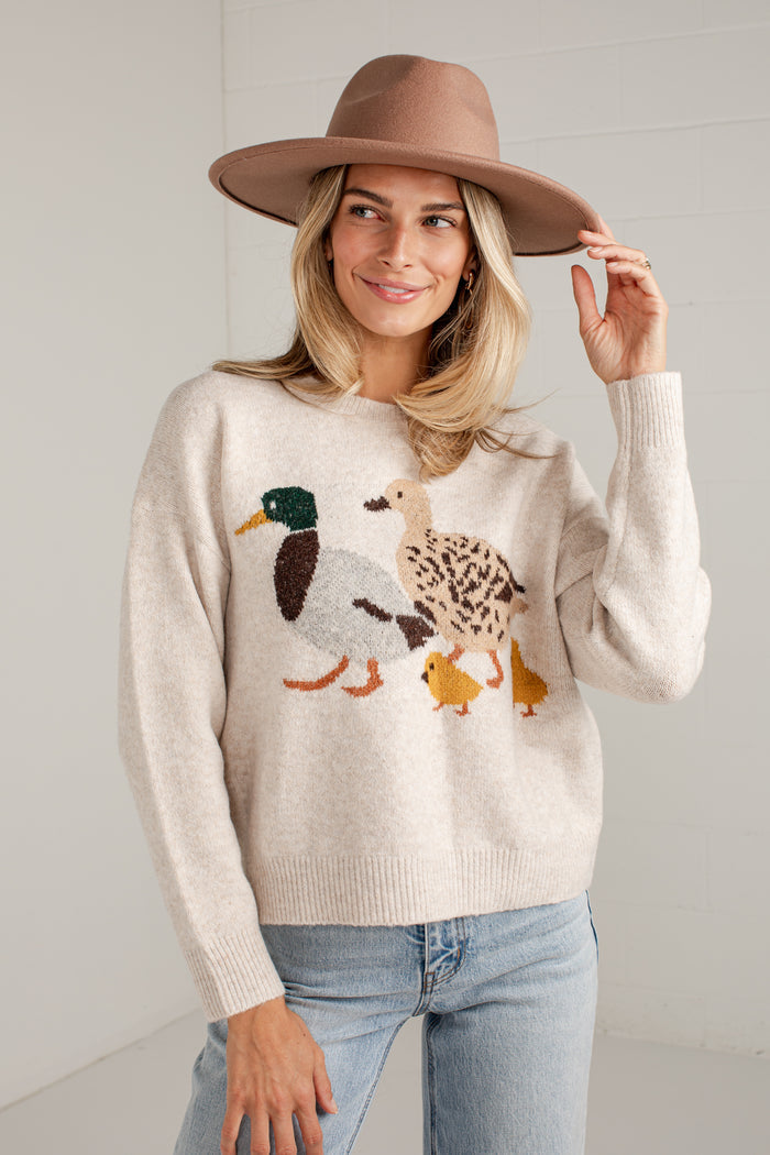 A woman wearing a cream knit sweater with a duck family motif, paired with light blue jeans and a brown wide-brim hat, standing against a white paneled wall.