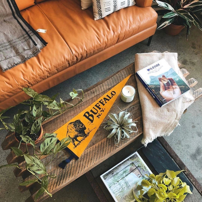 The Oxford Pennant Buffalo features a striking yellow wool felt design with a bold black illustration of a buffalo and the text "BUFFALO N.Y." in black. It is accented with black trim and two ribbon ties on the left, making it an ideal piece for Buffalo sports enthusiasts.