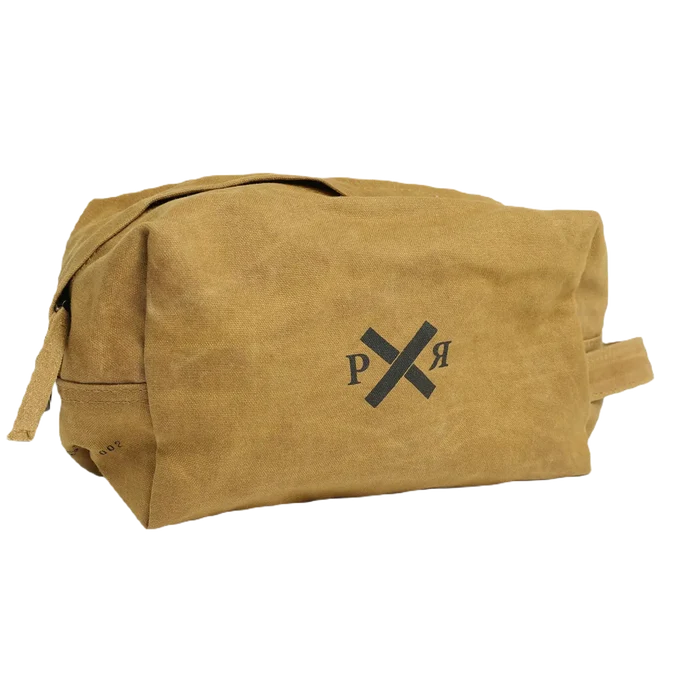 The Pony Rider Slow Road Waxed Canvas Toiletry Bag in Clay is a brown canvas grooming bag featuring a simple black logo with crossed lines and the letters "P" and "R." It includes a convenient handle zip closure and is made in India.