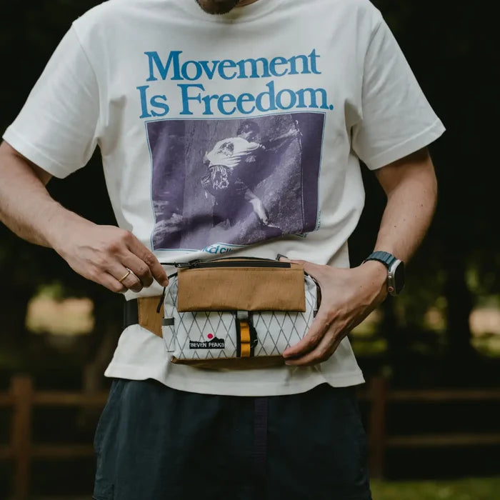 Person in a Movement Is Freedom shirt outdoors opens a brown Seven Peaks Fanny Pack, showcasing adventure gear style.