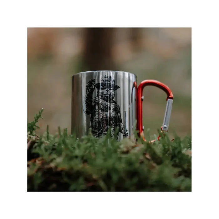 The Smokey Bear Carabiner Handle Mug - Waving Smokey is a silver mug that showcases an illustration of Smokey Bear wearing a ranger hat, featuring a vibrant red carabiner handle to promote wildfire prevention.