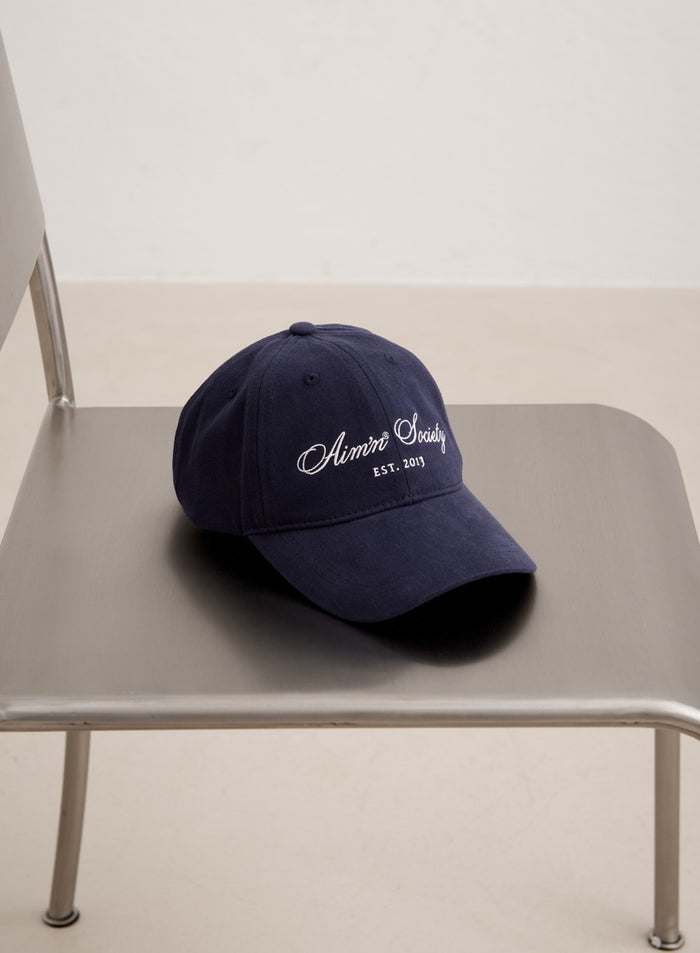 Tummansininen puuvillainen Society Cap valkoisella aim'n-kirjailulla harmaalla metallituolilla.