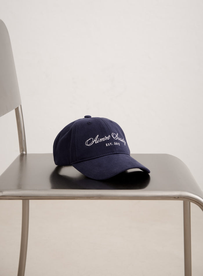 Tummansininen puuvillainen aim'n Society Cap valkoisella kirjailulla logolla minimalistisella metallituolilla.