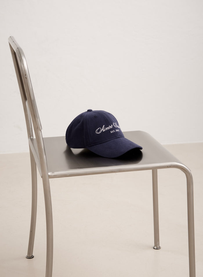 Tummansininen puuvillainen Society Cap valkoisilla kirjailluilla aim'n-logoilla hopeisella metallituolilla.
