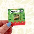 Blue-nailed hand displays Stacey McEvoy Caunt Deer Tile enamel pin (Aug 2026) on a vibrant green background.