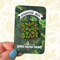 Hand displays Stacey McEvoy Caunt Green Man enamel pin on leafy card, perfect for nature, plant, and folklore lovers.