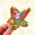 Hand displays a Stacey McEvoy Caunt Medieval Marginalia Harpy Vinyl Sticker, collectible art, August 2026 release.