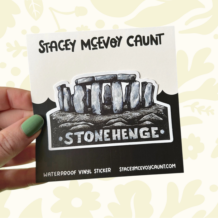 Hand holds Stacey McEvoy Caunt Stonehenge vinyl sticker; art supplies, pine cones in the background. Art sticker aesthetic.