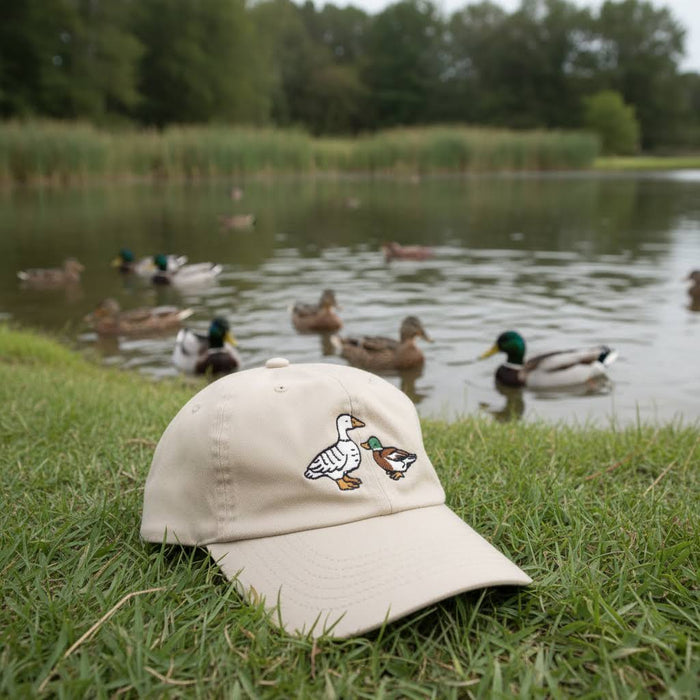 Forager Provisions LTD Stay Home Club beige cotton dad cap with embroidered duck design sitting on grassy pond bank.