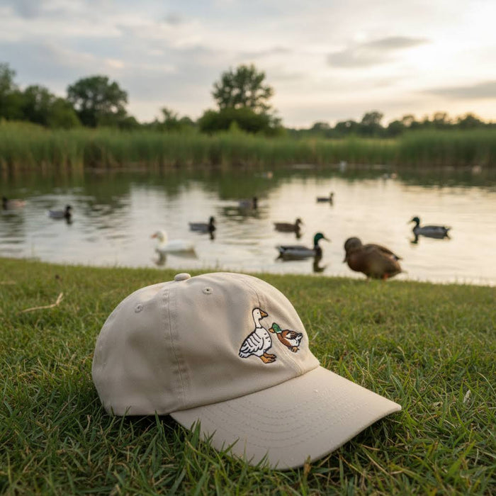 Forager Provisions LTD beige cotton dad cap with embroidered fowl design, resting on grass by a pond with ducks.